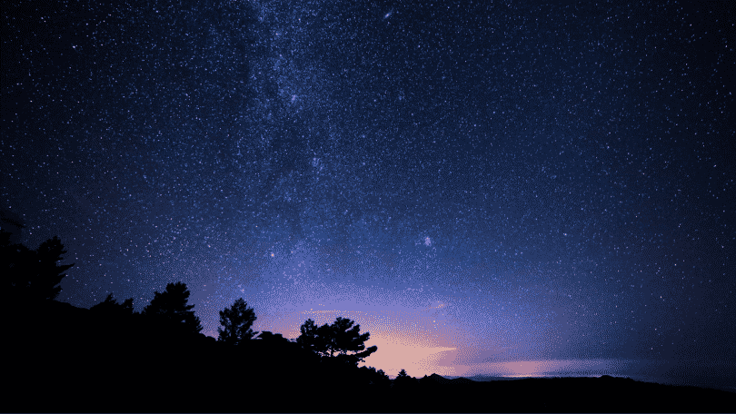 Imagem de um céu de noite, estrelado. Árvores ao fundo.