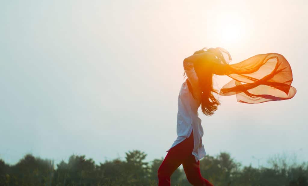 Mulher andando ao ar livre, segurando um lenço esvoaçante atrás da cabeça sob a luz do sol.