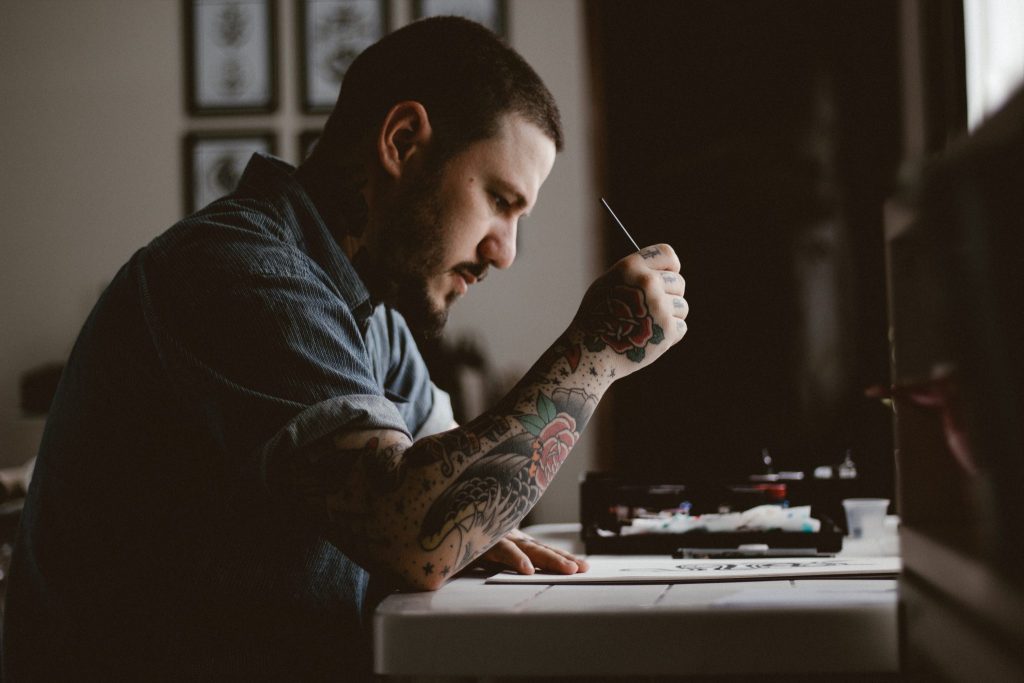 Homem com o braço tatuado, sentado em frente a uma mesinha. Ele está escrevendo algo em um papel.