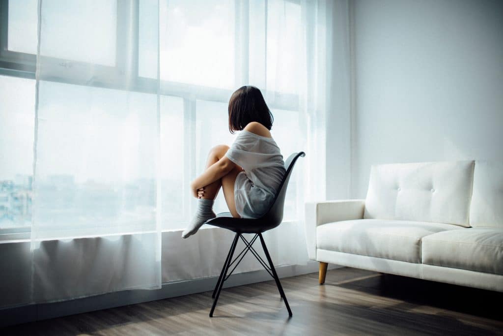 Menina sentada em uma cadeira, olhando pela janela, com as mãos segurando as pernas juntas ao peito.