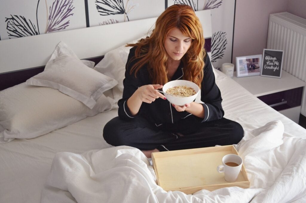 Mulher ruiva, sentada na cama, tomando café da manhã.