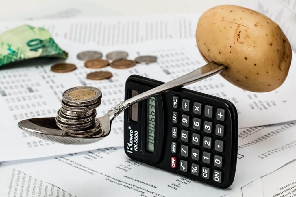 Uma colher em cima de uma calculadora equilibra uma batata de um lado e moedas de outro. Ao fundo, tem-se papeis, boletos e dinheiro em notas e moedas.