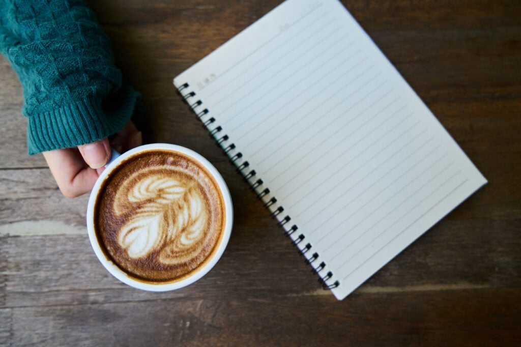 Pessoa segurando uma xícara de café, em uma mesa de madeira, ao lado de um caderno de anotações.