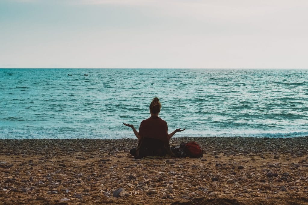 Silhueta de uma mulher de costas, na praia, meditando sentada na areia, de frente para o mar.