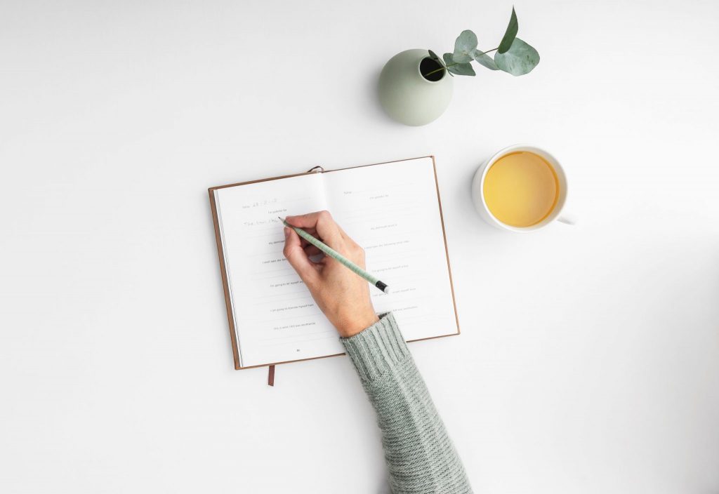 Foto da mão de uma menina escrevendo em um caderno. Ao lado há uma xícara com chá e uma plantinha.