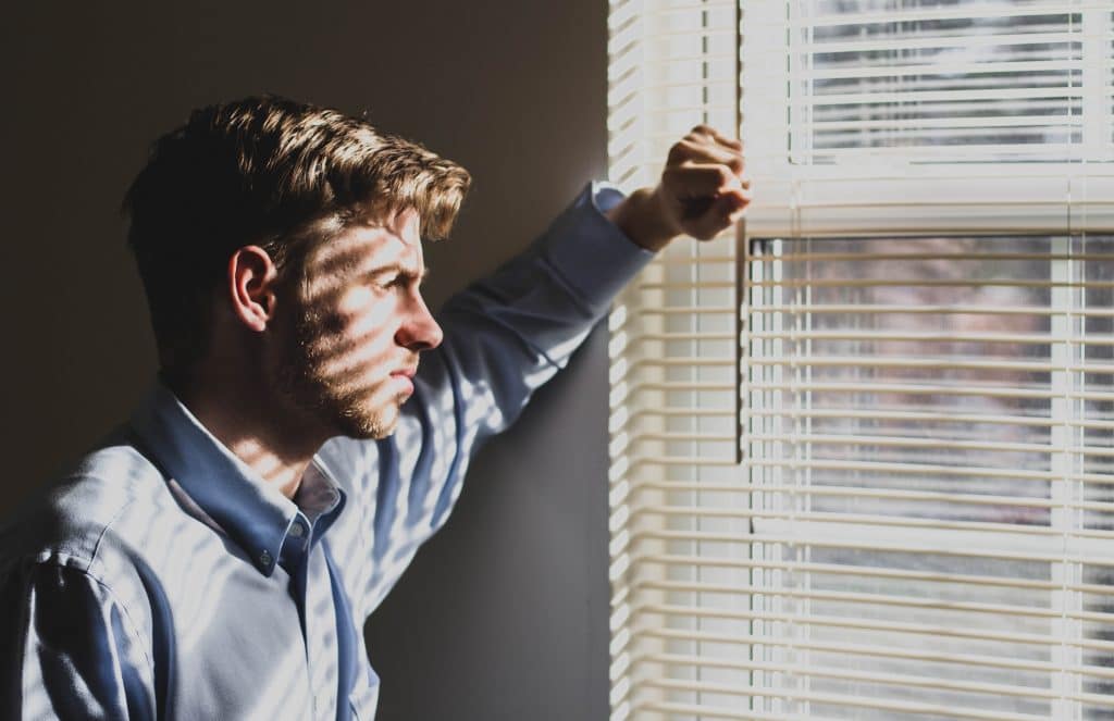 Homem com blusa social olhando pela janela, com a luz de fora refletindo em seu rosto.