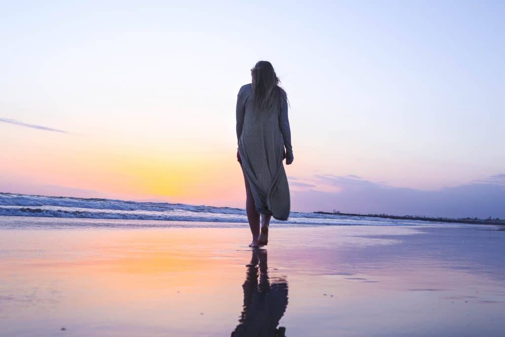 Mulher caminhando em praia sentido o mar com pôr-do-sol ao fundo