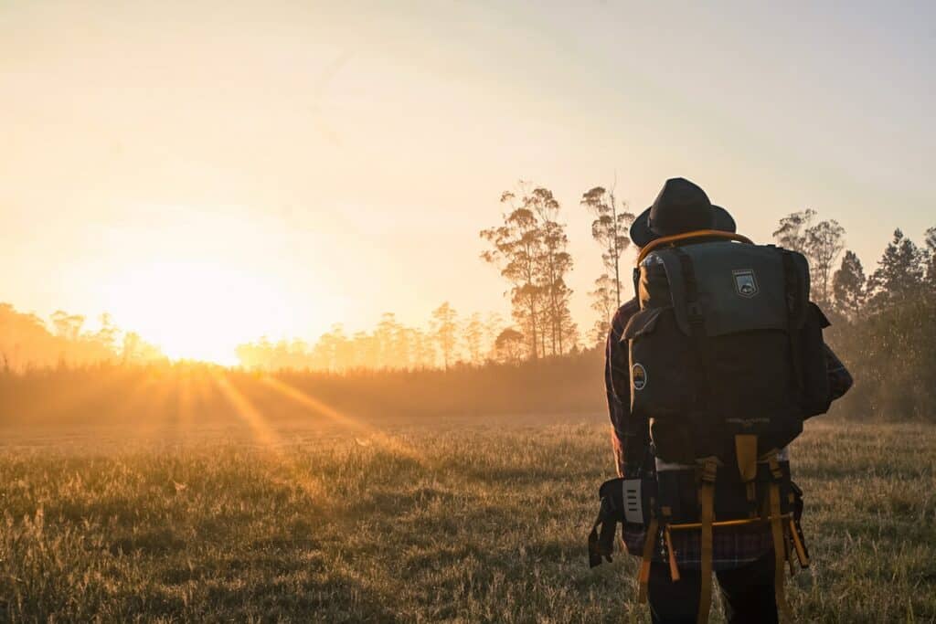 Pessoa viajante com chapéu e mochila nas costas olha para o nascer do sol.