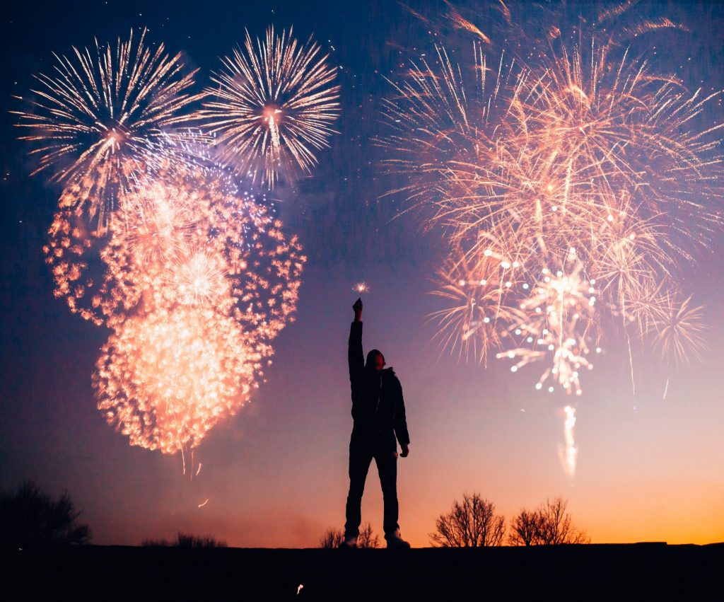 Homem segurando vela estrelinha com fogos artificiais ao fundo estourando