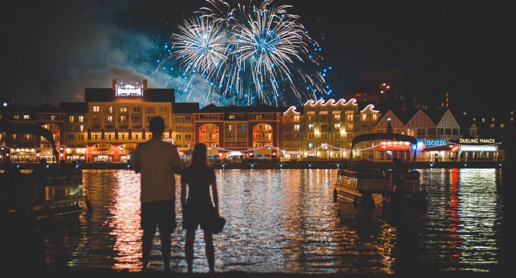 Casal de costas em frente a um rio, admirando os fogos de artifício sendo estourados no céu noturno.