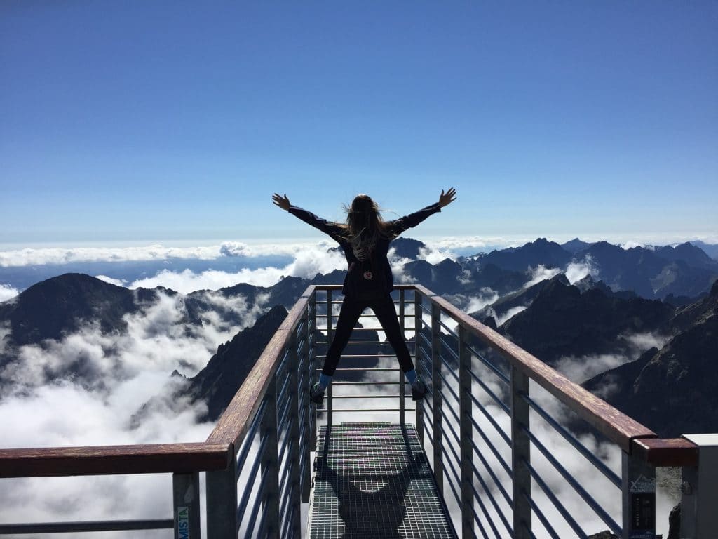 Mulher de braços abertos em ponte com montanhas e céu azul ao fundo 