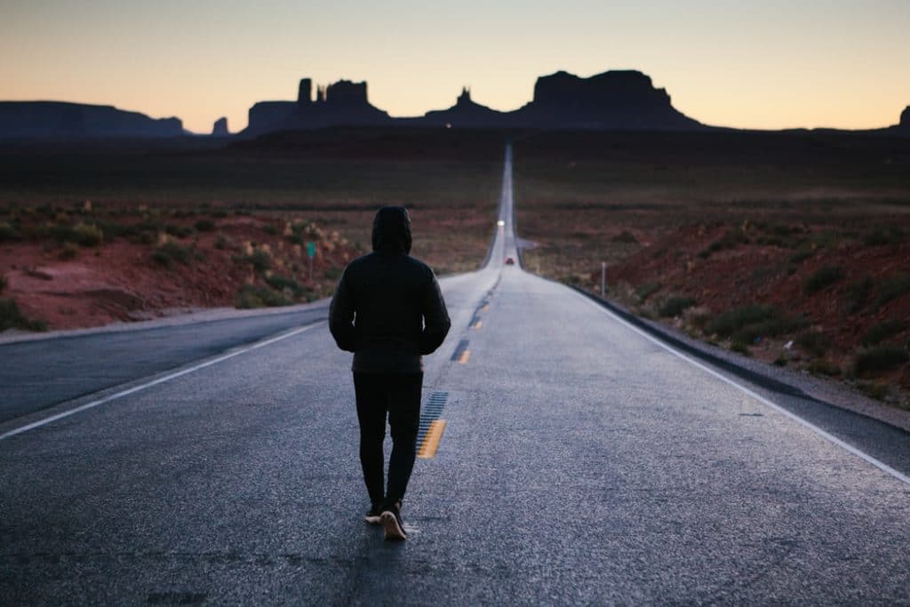 Homem de costas, de calça e capuz, andando sozinho no meio de uma estrada, no fim da tarde.