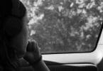 Foto em preto e branco de mulher jovem olhando pela janela do carro. Fora vemos árvores e mata fechada. A mulher apoia sua boca na mão esquerda, de forma pensativa.