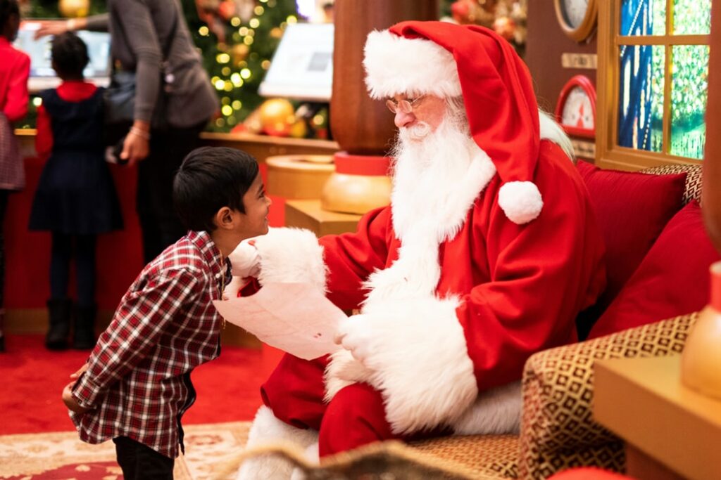 Menino pequeno em frente de um homem vestido como Papai Noel, que segura uma cartinha enquanto olha para a criança.