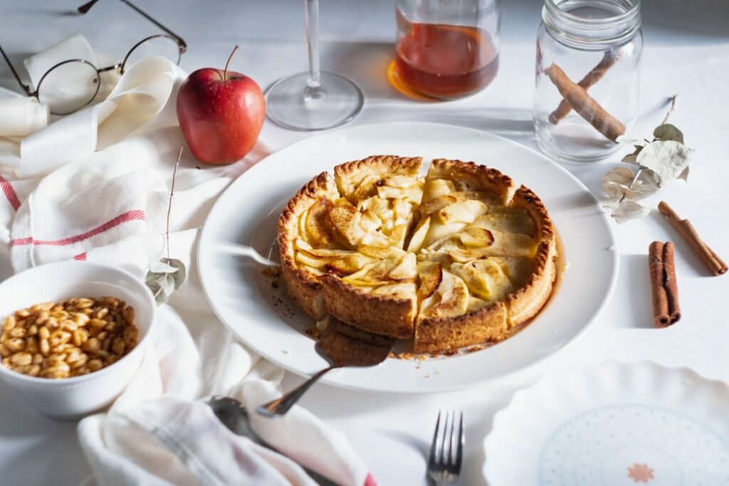 Torta de maçã em prato branco, com cortador de bolo embaixo de uma fatia. Ao redor, na mesa, um pote com paus de cnaela, um pote com amendoins , uma maçã, talheres e mel.