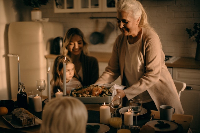 Mulher idosa colocando uma forma com um chester assado na mesa de jantar. Uma mulher com uma criança no colo sorri. Várias taças, copos e velas acesas estão na mesa. 