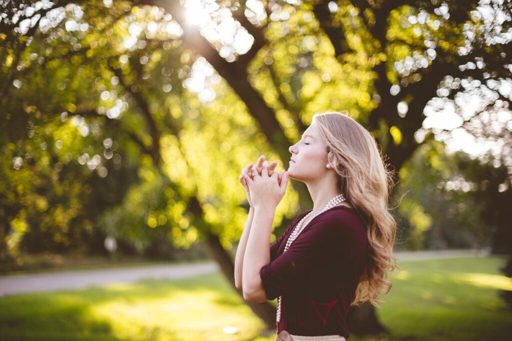 Mulher loira, em um parque, com as mãos jutas em frente ao queixo e os olhos fechados, orando.