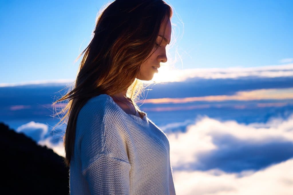 Mulher em pé, de perfil, olhando para baixo. Ao fundo, há uma paisagem com nuvens e a luz do sol.