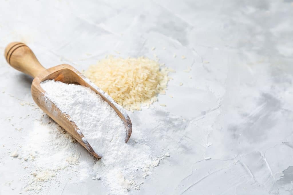 Farinha esparramada sobre uma mesa enfarinhada. Sobre a mesa ainda temos uma colher grande de madeira cheia de farinha de trigo e ao lado uma pordão de arroz. Direitos Autorais:  Oleksandra Naumenko.