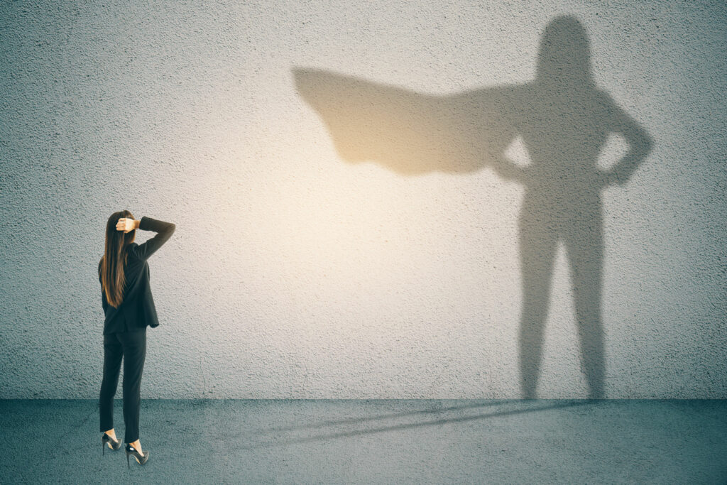 Mulher com traje social, olhando para uma parede branca. Sua sombra a representa como uma heroína, vestindo uma capa, com as mãos na cintura.
