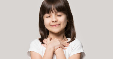 Menina com as duas mãos no peito, olhos fechados e esboçando um sorriso.