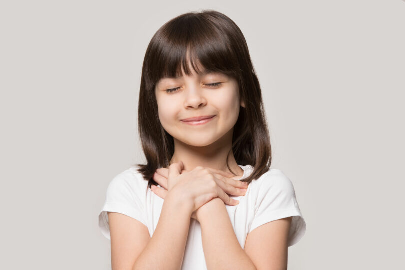 Menina com as duas mãos no peito, olhos fechados e esboçando um sorriso.