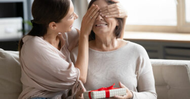 Mulher jovem cobrindo os olhos de uma mulher mais velha, que segura um presente.