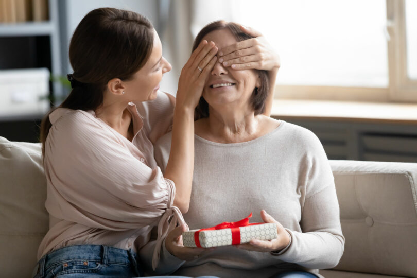 Mulher jovem cobrindo os olhos de uma mulher mais velha, que segura um presente.