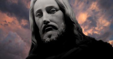 Rosto de estátua de Jesus Cristo, com nuvens escuras encobrindo um céu alaranjado ao fundo.