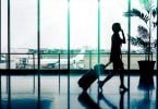 Silhueta de mulher andando em aeroporto, puxando uma mala de rodinhas com uma bolsa apoiada em cima, enquanto fala no celular. Ela está em frente a um vidro onde é possível ver um avião e a pista de pouso e decolagem.