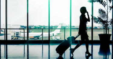 Silhueta de mulher andando em aeroporto, puxando uma mala de rodinhas com uma bolsa apoiada em cima, enquanto fala no celular. Ela está em frente a um vidro onde é possível ver um avião e a pista de pouso e decolagem.