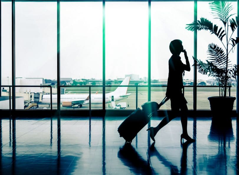 Silhueta de mulher andando em aeroporto, puxando uma mala de rodinhas com uma bolsa apoiada em cima, enquanto fala no celular. Ela está em frente a um vidro onde é possível ver um avião e a pista de pouso e decolagem.