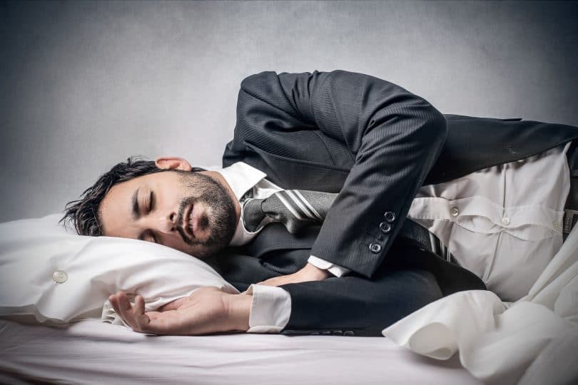 Homem deitado na cama, dormindo, com roupas sociais de trabalho.