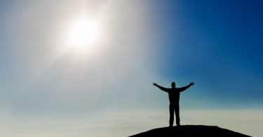 Silhueta de homem de costas com braços abertos no topo de montanha com céu azul e sol ao fundo