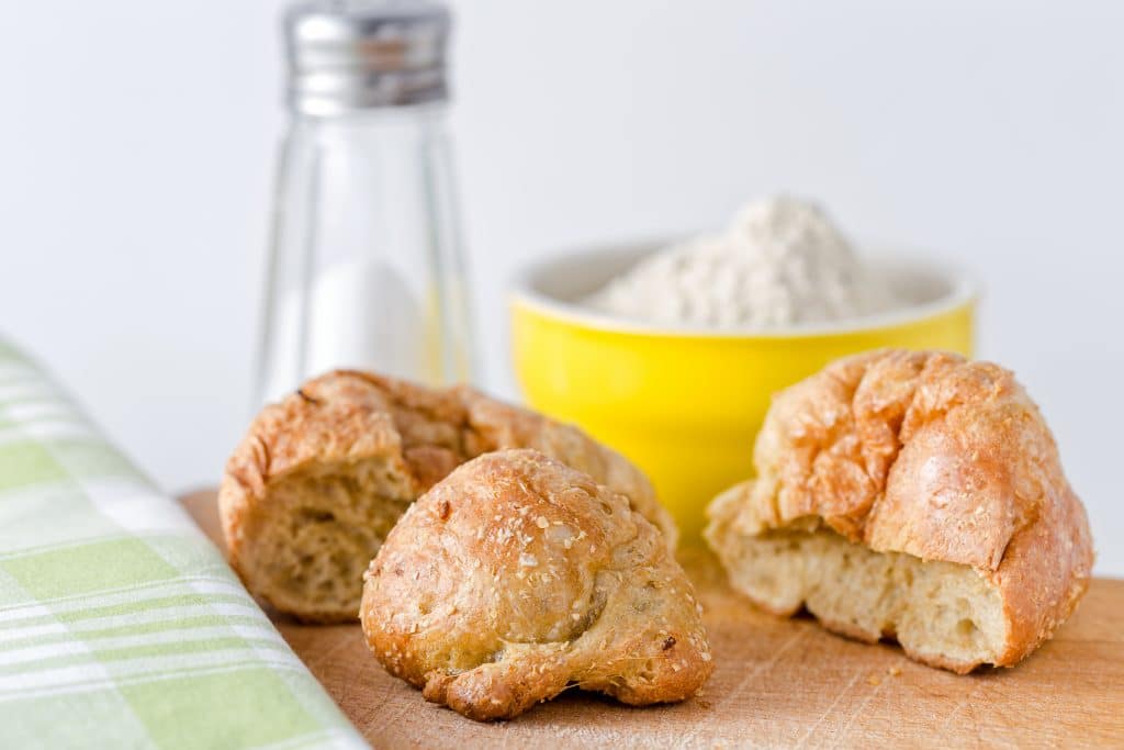 Três pedaços de pão assados, sobre uma mesa de madeira. Ao lado um pote amarelo cheio de farinha branca e um toalha de mesa xadrex nas cores branco e verde. Direitos autorais : Massimiliano Ranauro 
