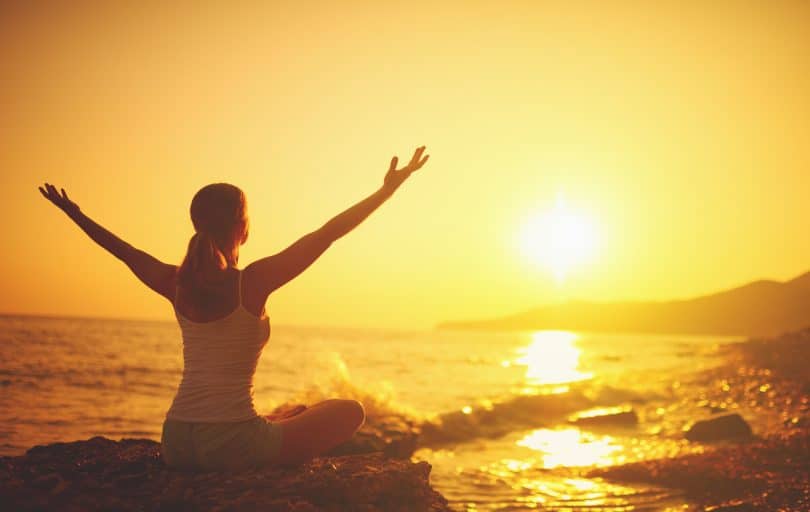 Mulher sentada em pedra numa praia, com os braços abertos, olhando para o pôr do sol.