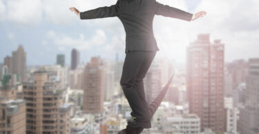 Homem com terno e calça social, se equilibrando em corda, que cruza uma cidade inteira. Ele se equilibra com os braços abertos. Ao fundo, a cidade, com diversos prédios.