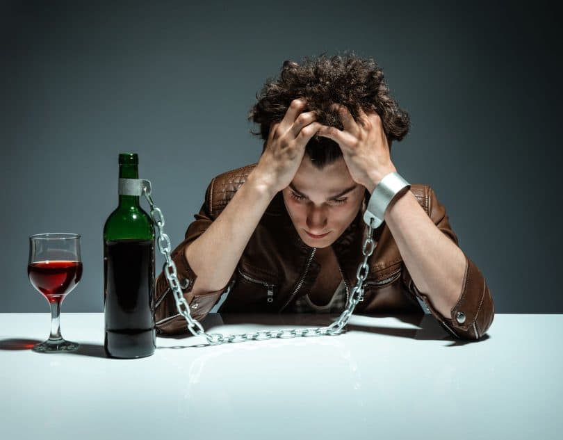 Homem com os cotovelos apoiados numa mesa, e sua testa está apoiada em suas mãos. Grilhões prendem seu braço esquerdo a uma garrafa de vinho, que está em cima da mesa, com uma taça de vinho cheia ao lado.