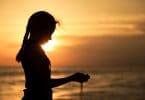 Silhueta de menina em praia, com o pôr do sol ao fundo, segurando areia com as duas mãos, e a areia escapando entre seus dedos.