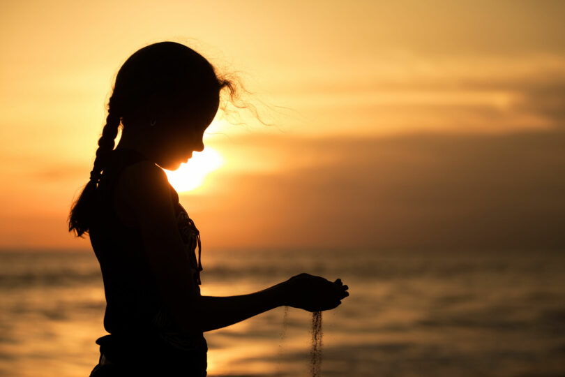 Silhueta de menina em praia, com o pôr do sol ao fundo, segurando areia com as duas mãos, e a areia escapando entre seus dedos.