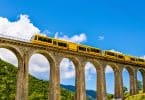 Trem amarelo passando por uma alta ponte. Ao fundo, montanhas arborizadas e um céu bem azul.