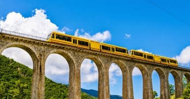 Trem amarelo passando por uma alta ponte. Ao fundo, montanhas arborizadas e um céu bem azul.