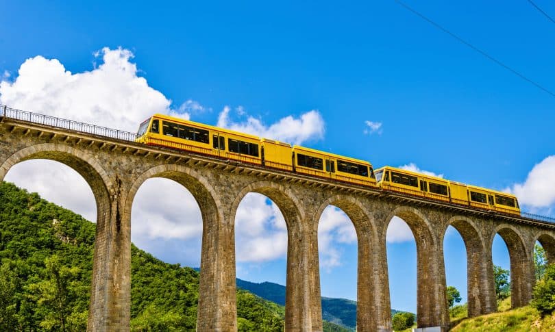 Trem amarelo passando por uma alta ponte. Ao fundo, montanhas arborizadas e um céu bem azul.