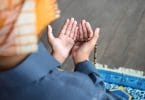 Pessoa com turbante ajoelhada em um tapete, com as mãos estendidas em sinal de oração.