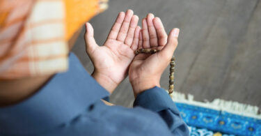 Pessoa com turbante ajoelhada em um tapete, com as mãos estendidas em sinal de oração.