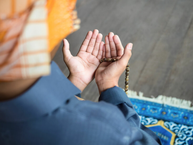 Pessoa com turbante ajoelhada em um tapete, com as mãos estendidas em sinal de oração.