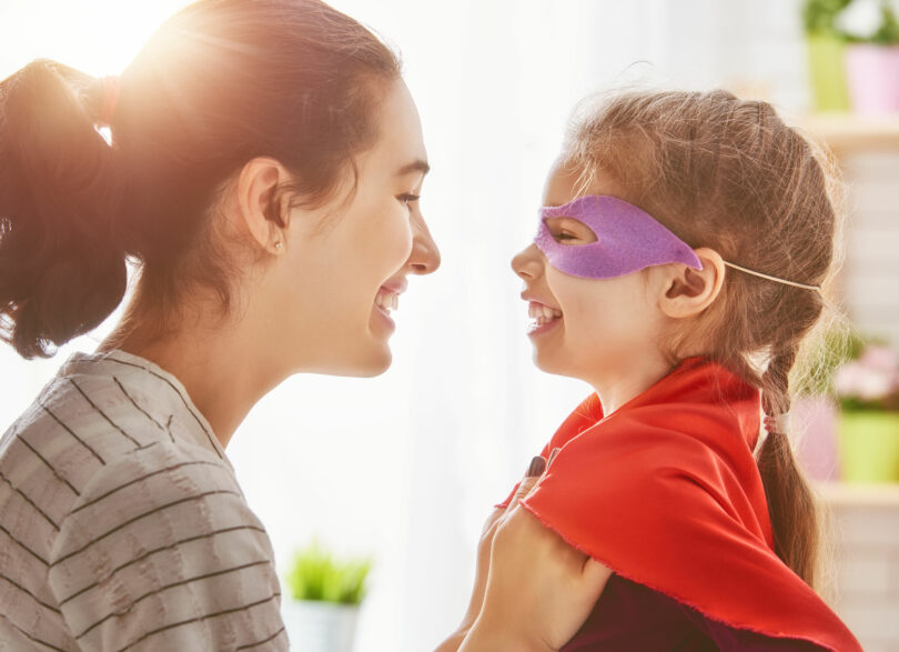 Mãe arrumando capa de super-herói da filha que está sorrindo