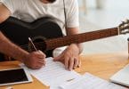Homem sentado em uma mesa com um violão sobre o colo enquanto escreve músicas em uma folha.