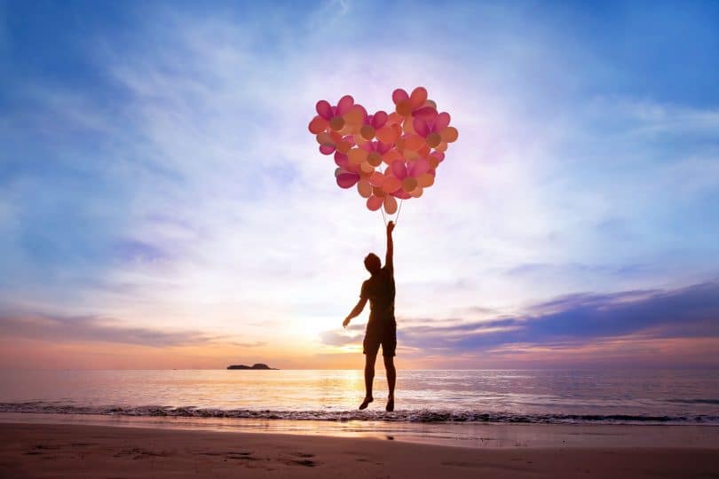 Silhueta de pessoa segurando balões formando um coração na praia