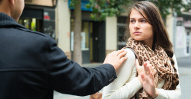 Homem seguindo mulher na rua e encostando em seu ombro. A mulher olha com para o homem incomodada, e estende sua mão para ele se distanciar.